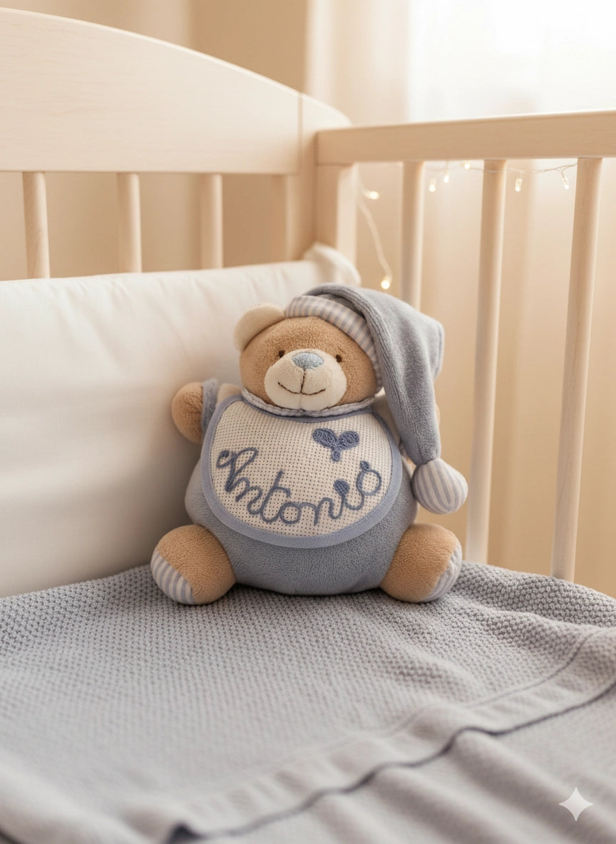 Orsetto peluche per neonati con pigiamino e cappellino a righe. Il peluche ha un bavaglino bianco in tela Aida per ricamo.